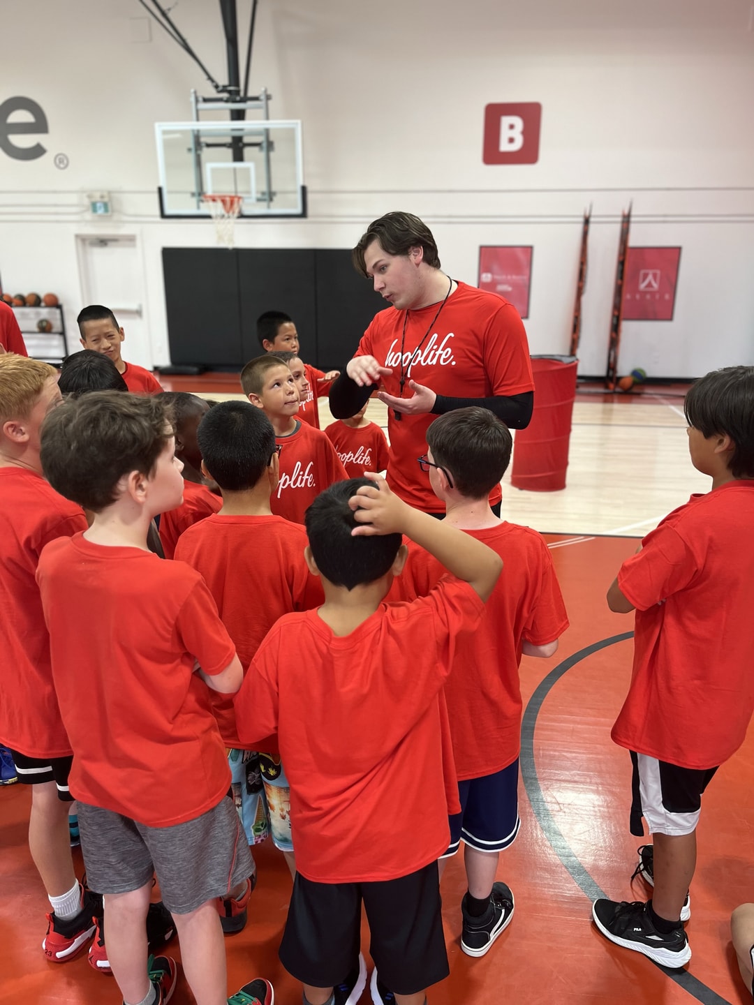 A HoopLife coach in a huddle with a group of kids, partner signage on the wall behind